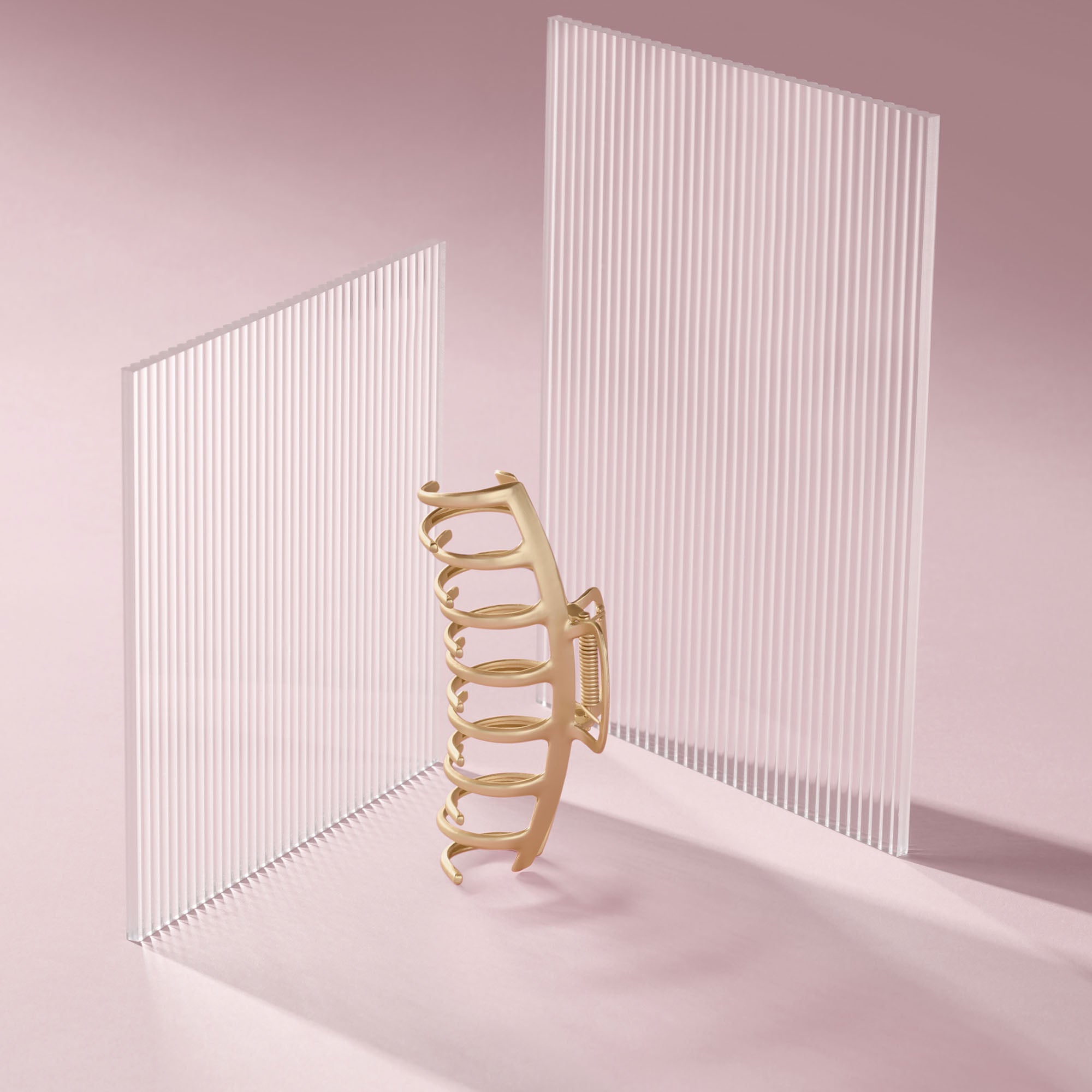 The Hair Edit Gilded Timeless Claw Clip stands upright between two translucent white slatted panels on a soft pink studio background.5