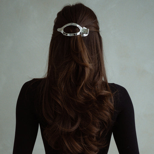 A woman viewed from the back wearing a shiny silver metal hair claw clip holding up her dark brown hair, against a plain light gray background.