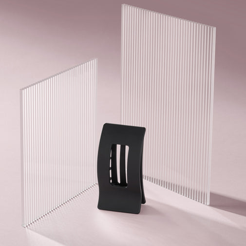 A black rectangle flat lay claw clip standing up, displayed in front of two tall white slatted panels on a soft pink studio background.