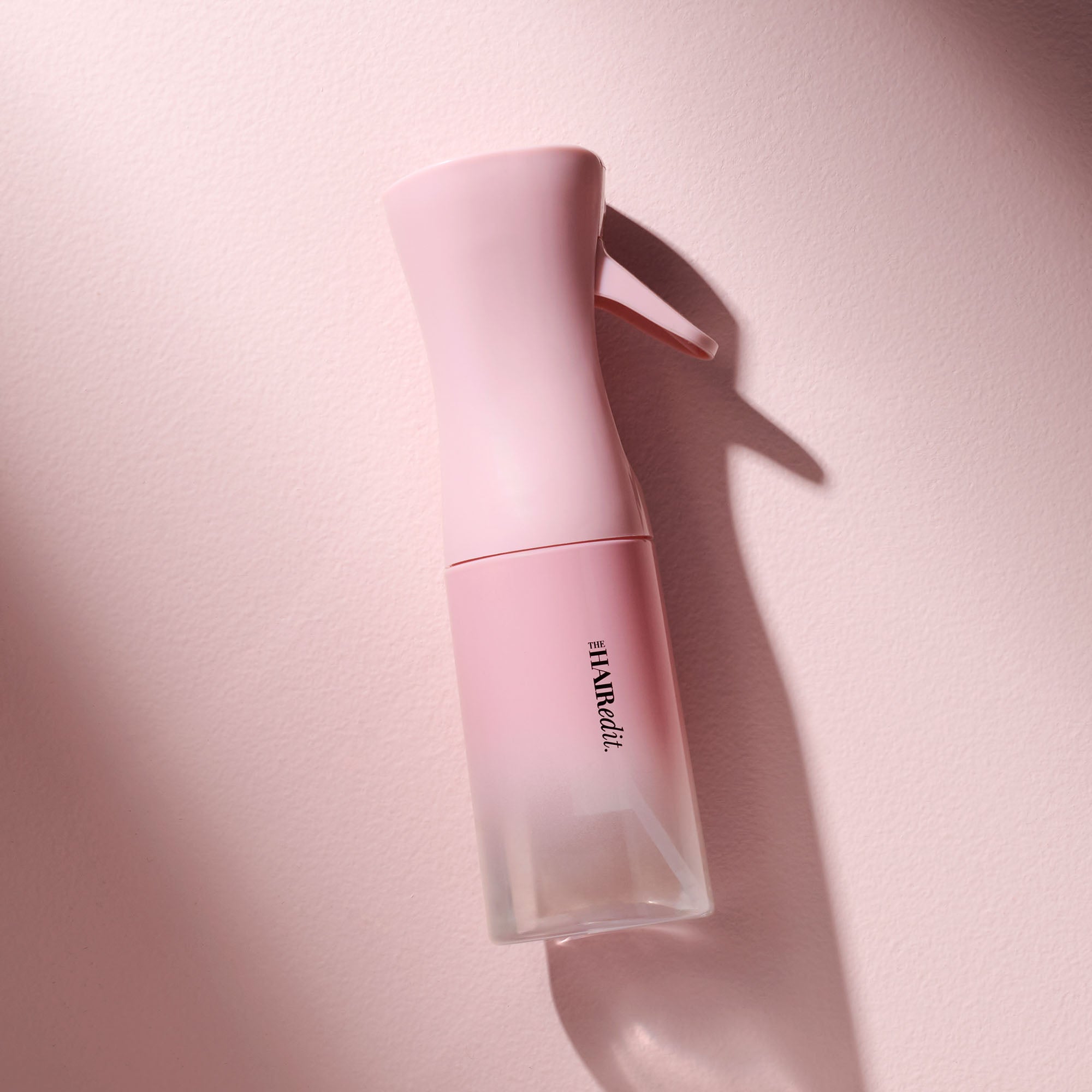 A pink spray bottle with a clear lower section and black vertical text reading 'THE HAIR EDIT', photographed on a soft pink background with a strong shadow.6