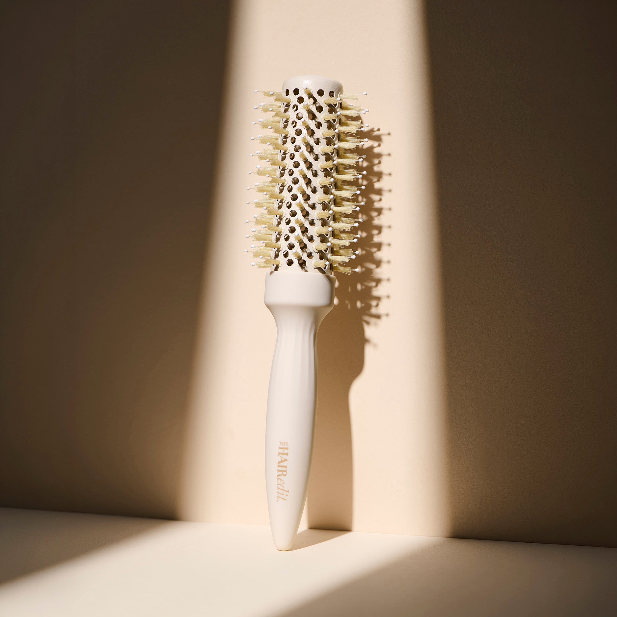 A white round hairbrush with cream-colored bristles and a white handle, standing upright against a beige background with dramatic diagonal shadows.5