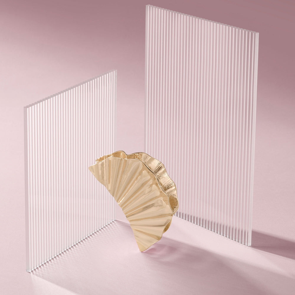 A gold pleated fan-shaped claw clip stands in front of two translucent white slatted panels on a soft pink studio background.6