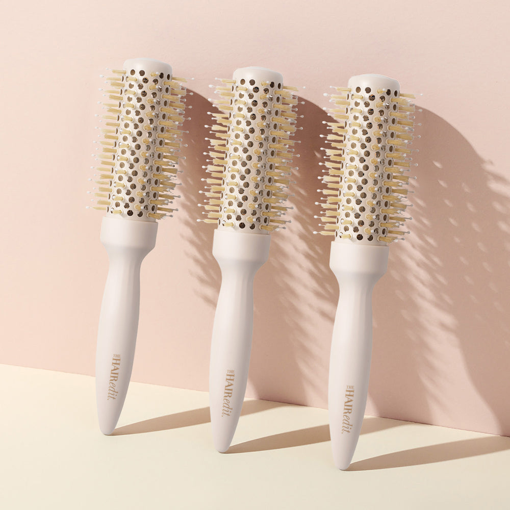 Three round hairbrushes with white handles and beige bristles, arranged side by side on a cream surface against a soft blush pink background.6