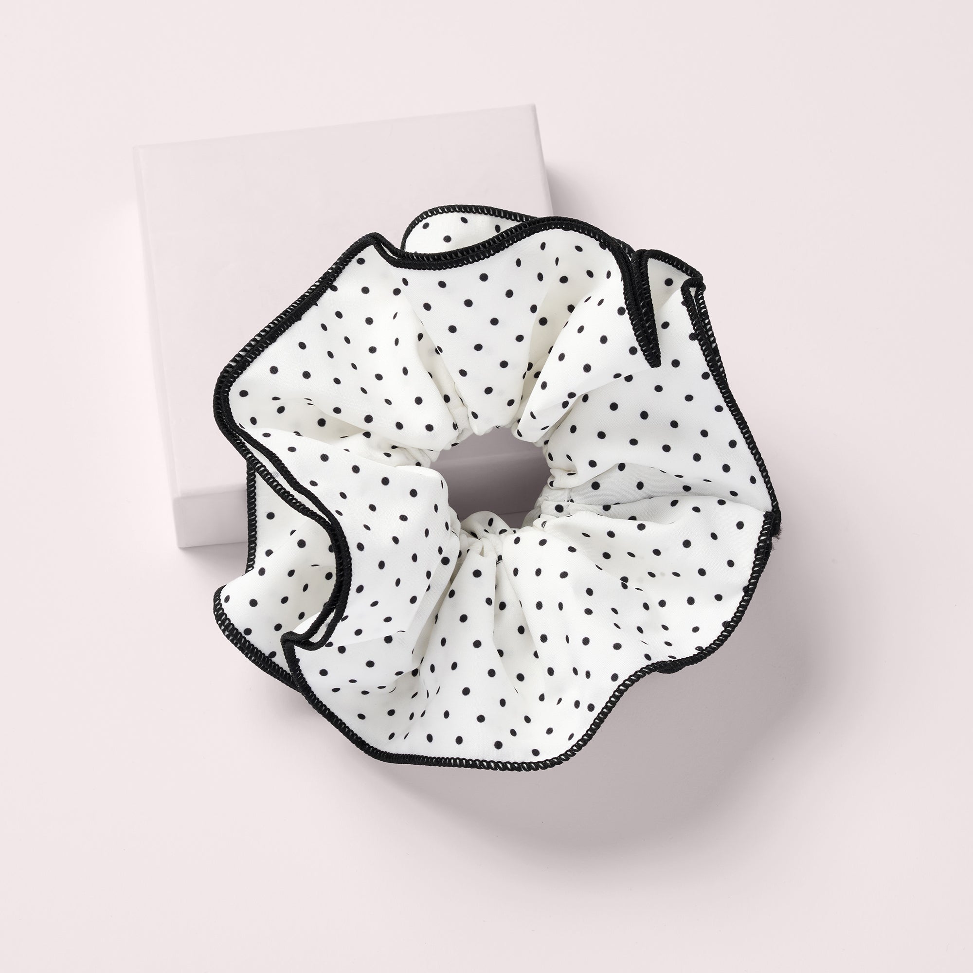 A white fabric scrunchie with black polka dots and black stitched trim, displayed on a light pink background with a white square box behind it.9