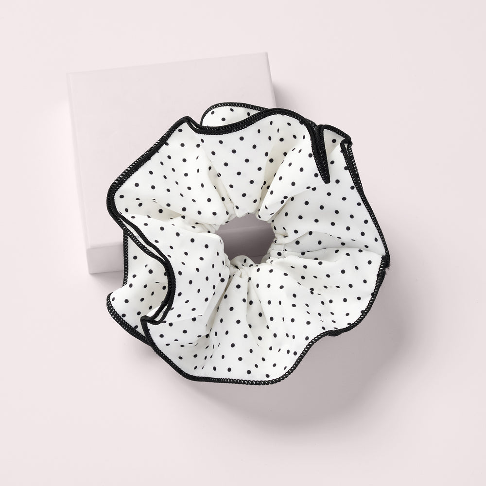 A white fabric scrunchie with black polka dots and black stitched trim, displayed on a light pink background with a white square box behind it.9