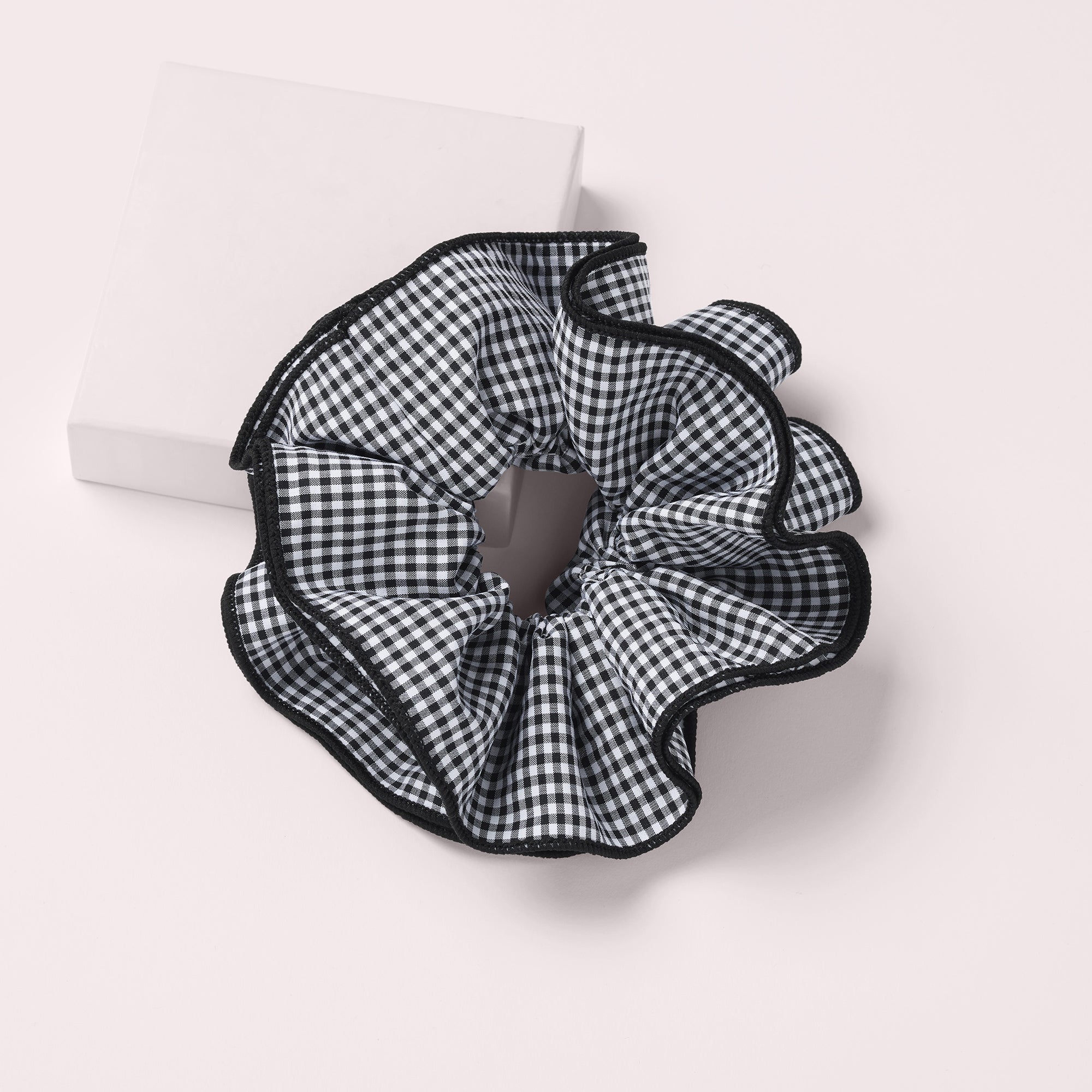 A black and white gingham patterned scrunchie with a black ruffled trim, displayed on a light beige background next to a white rectangular box.8