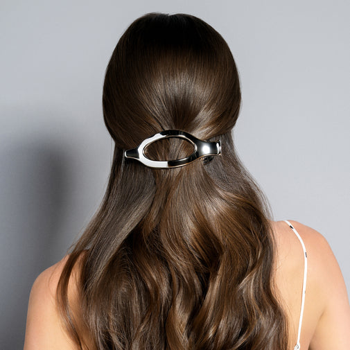 A woman viewed from the back with long, wavy brown hair and a shiny silver oval hair claw clip holding the hair at the crown, against a plain light gray background.