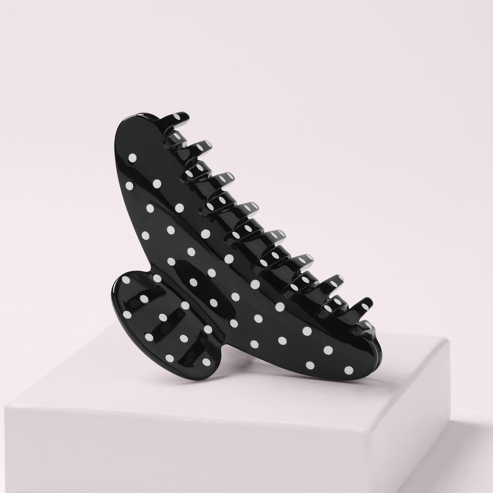 A glossy black claw hair clip with white polka dots, displayed on a white pedestal against a light beige background.8
