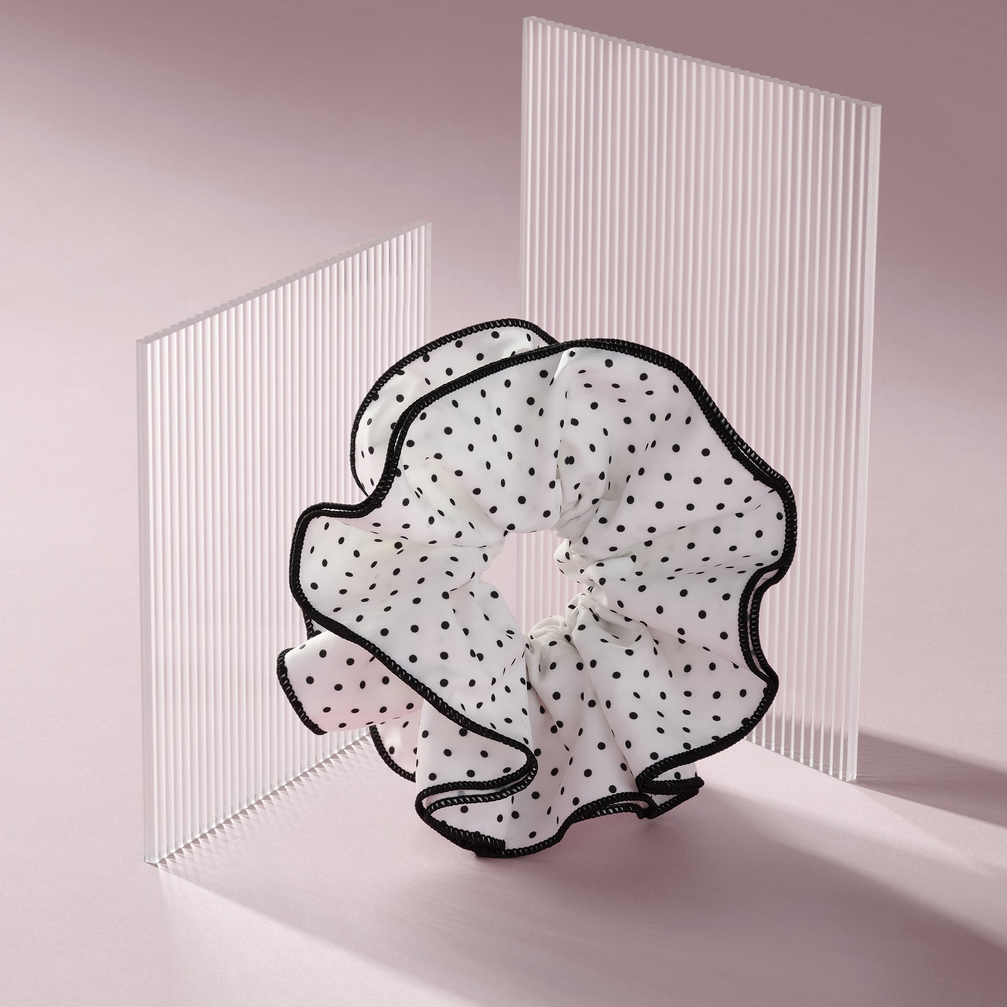 A sheer white ruffled hair scrunchie with black polka dots and black trim, displayed on a minimalist pink studio background with translucent vertical panels.10