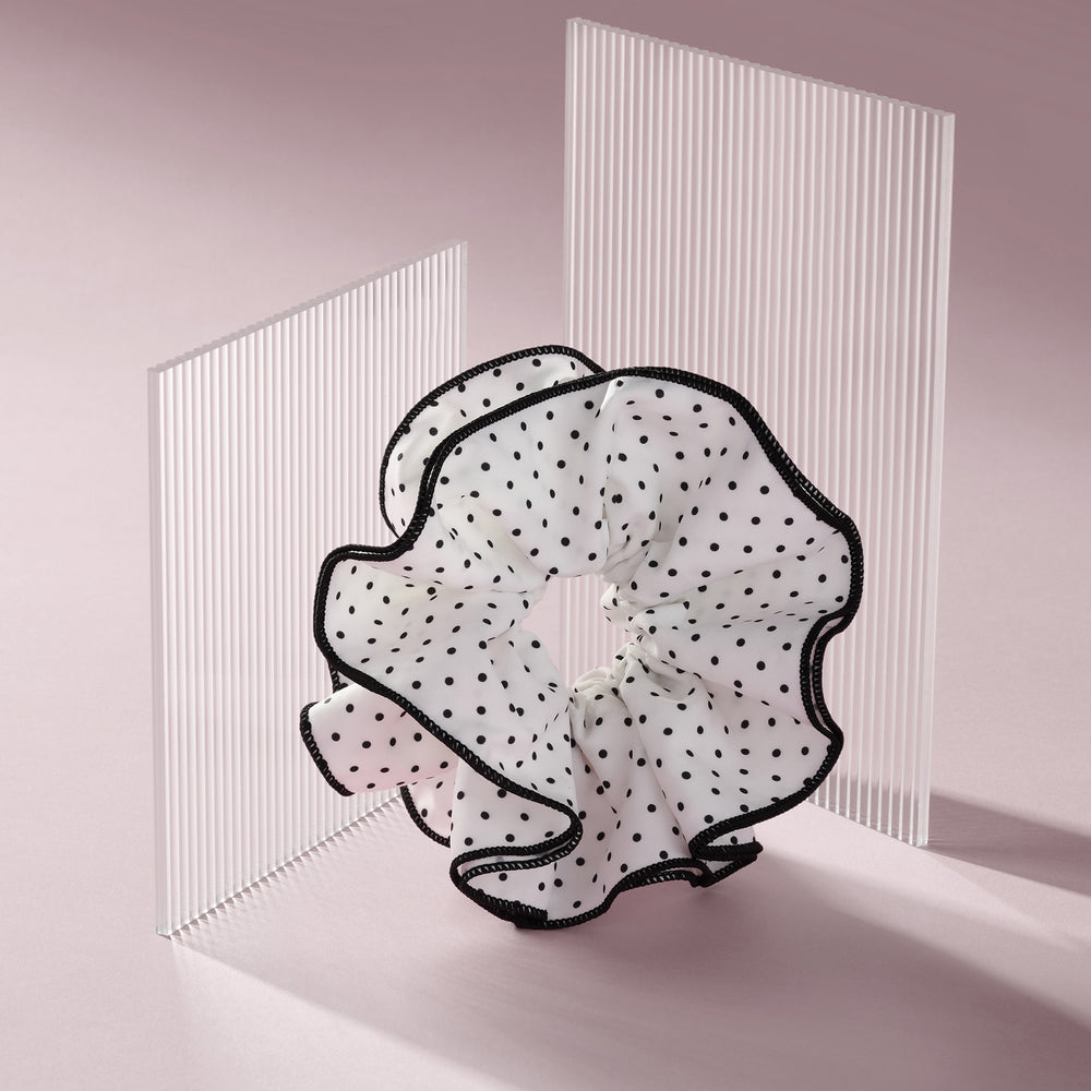 A sheer white ruffled hair scrunchie with black polka dots and black trim, displayed on a minimalist pink studio background with translucent vertical panels.10