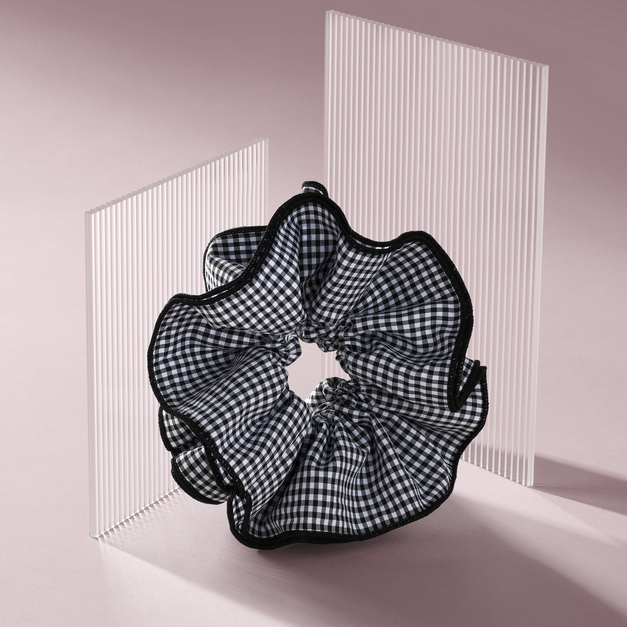 A black and white gingham check scrunchie with black ruffled trim, displayed on a minimalist pink and white studio set.9