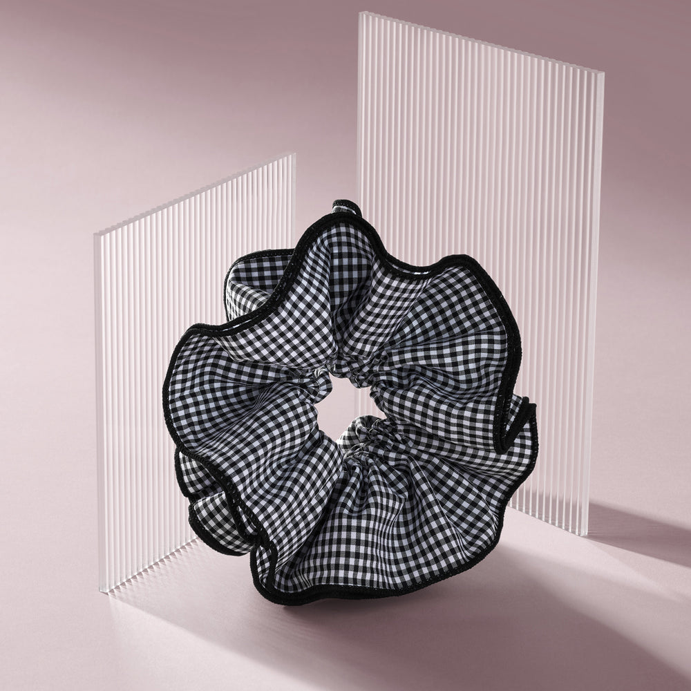 A black and white gingham check scrunchie with black ruffled trim, displayed on a minimalist pink and white studio set.9