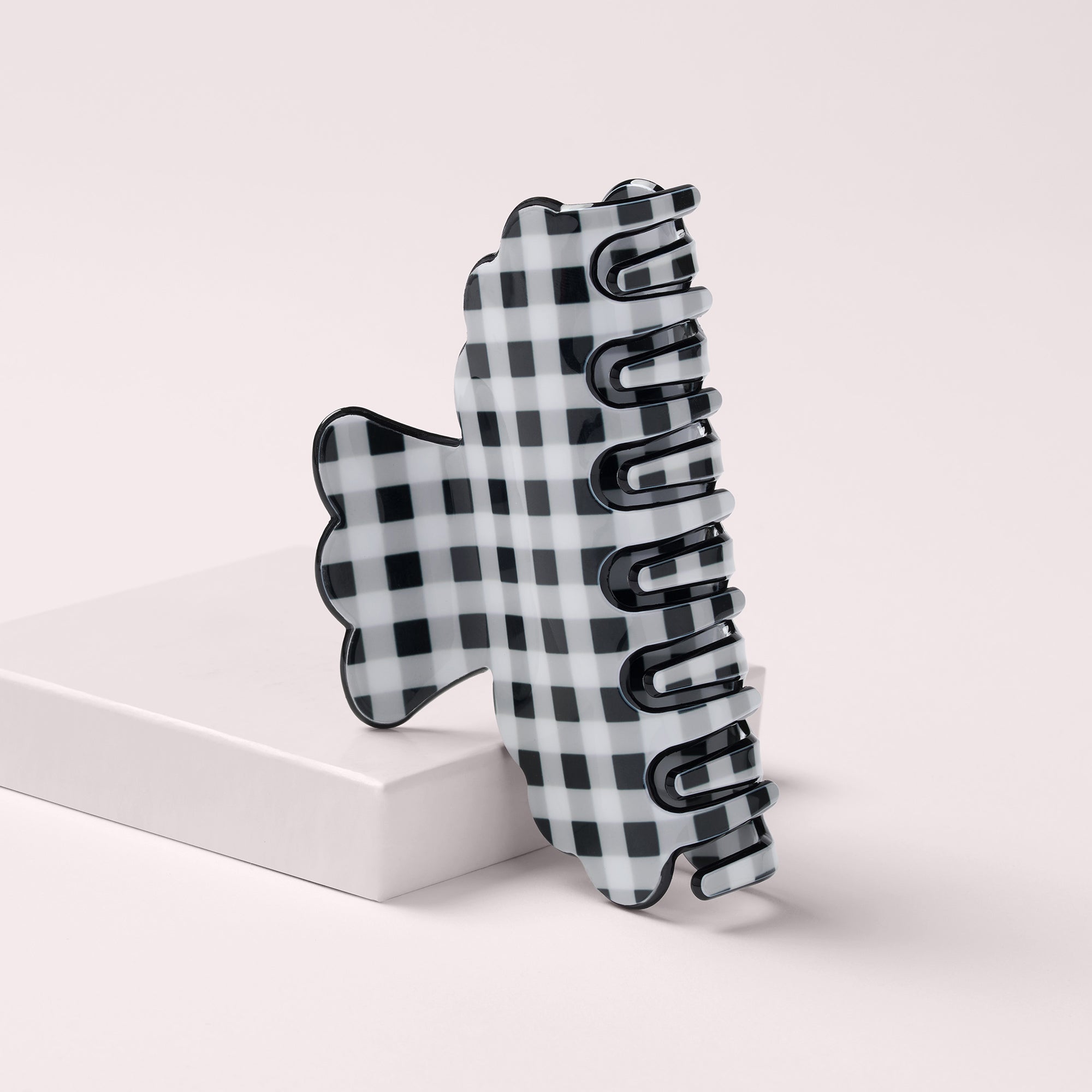 A black and white checkered claw hair clip standing upright on a white rectangular block against a light beige background.8