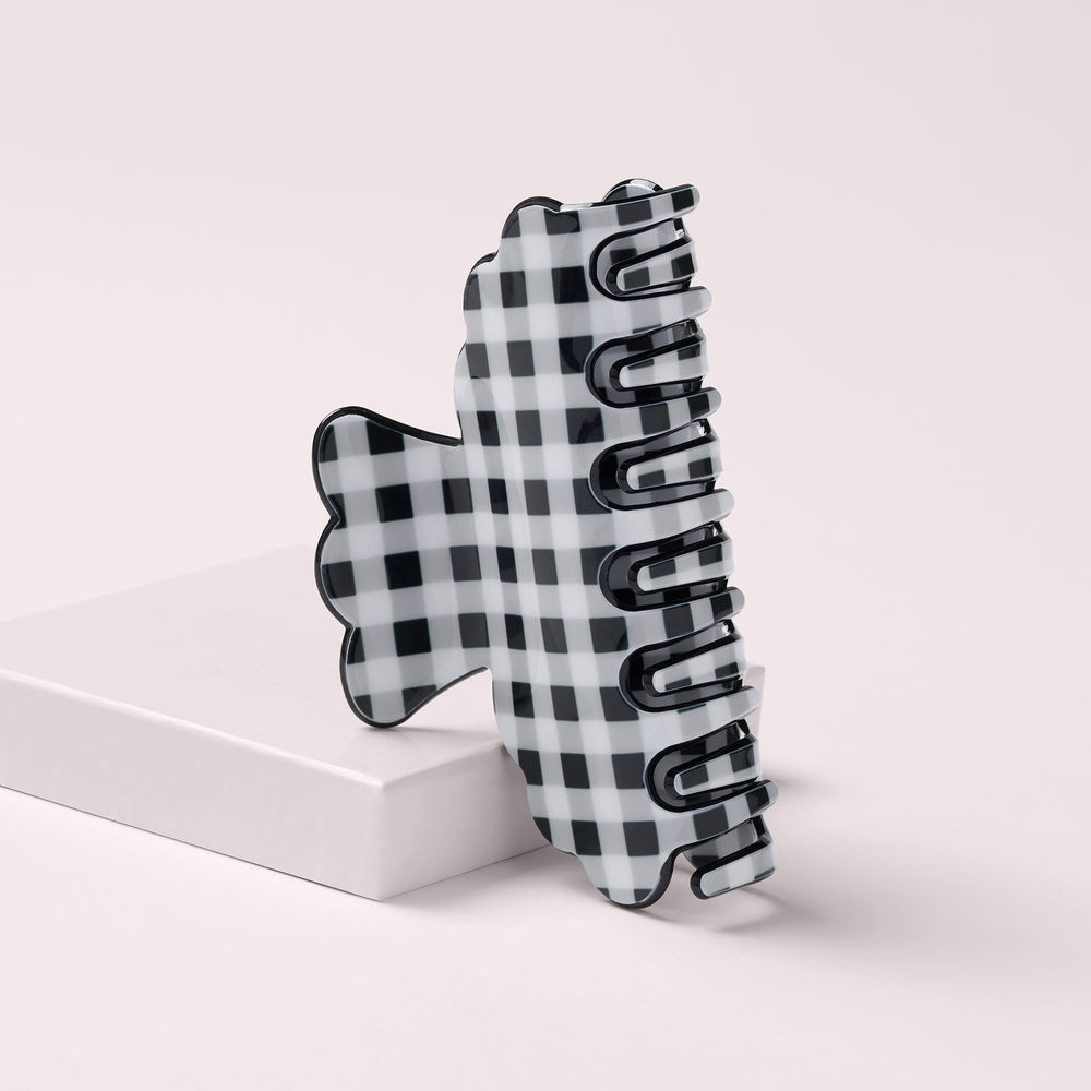 A black and white checkered claw hair clip standing upright on a white rectangular block against a light beige background.8