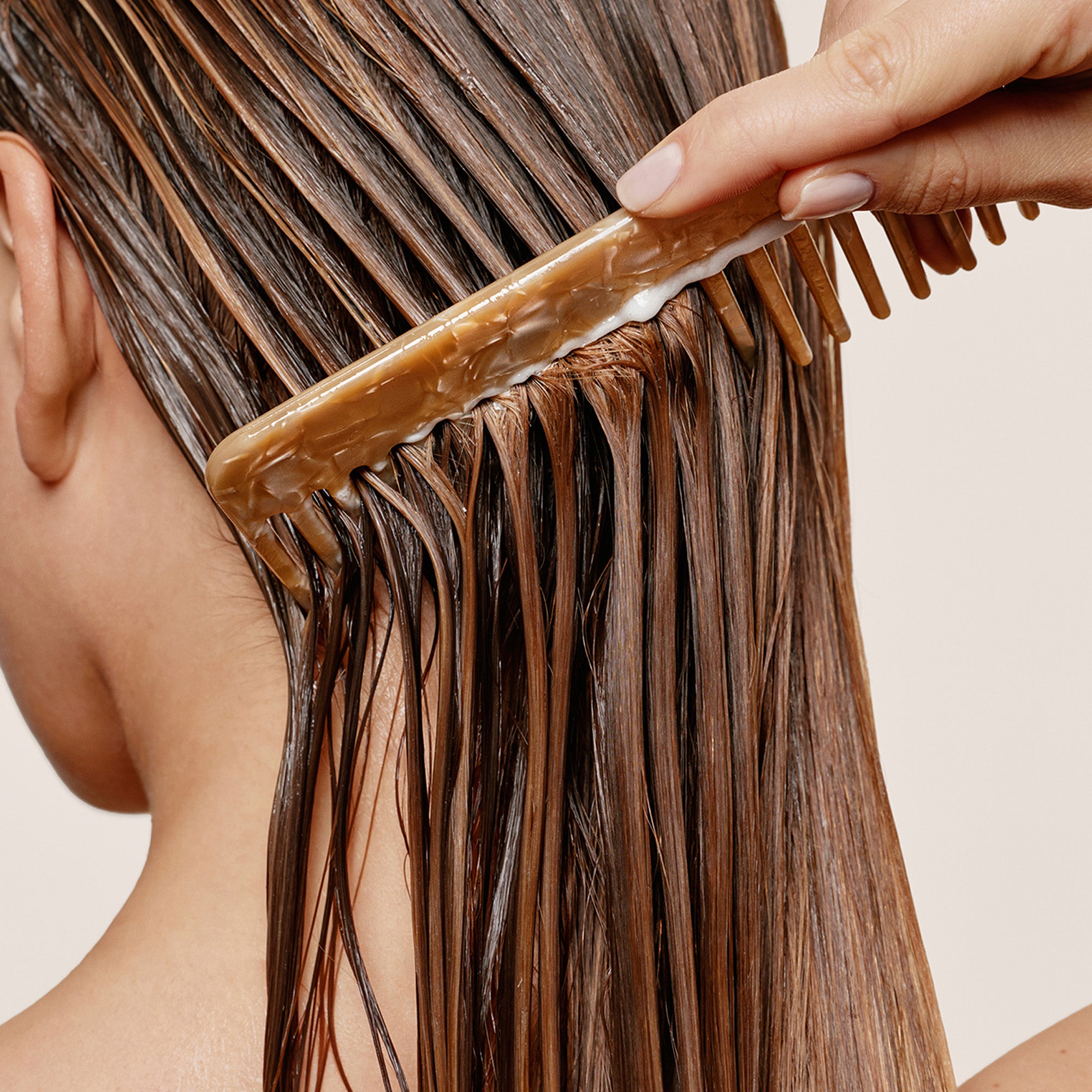 A close-up of a person's long, dark brown hair being gently combed with a translucent amber-brown hair tool held by a hand against a plain light beige background.7