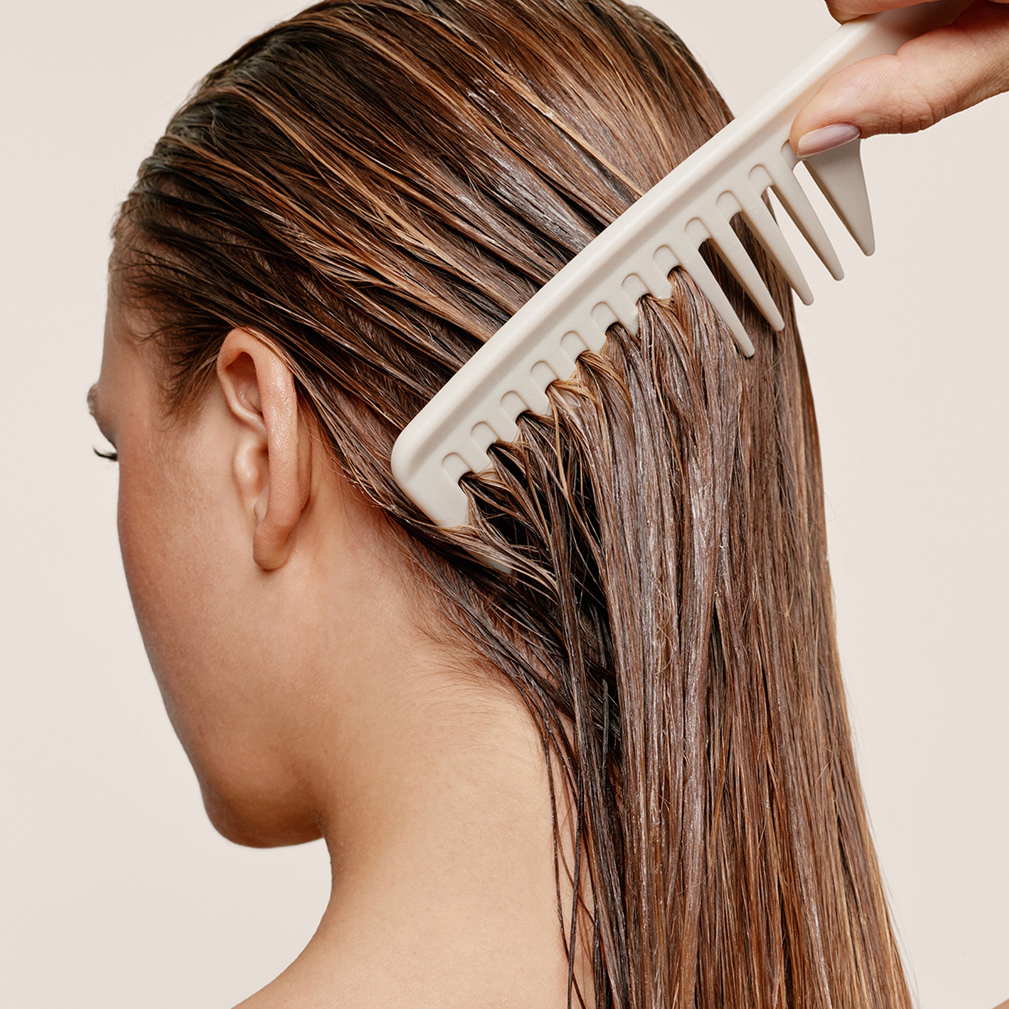 A close-up side view of a person with long, wet brown hair being combed through the hairline with a wide-tooth white comb. A hand is visible holding the comb, and the background is a clean light beige studio backdrop.7