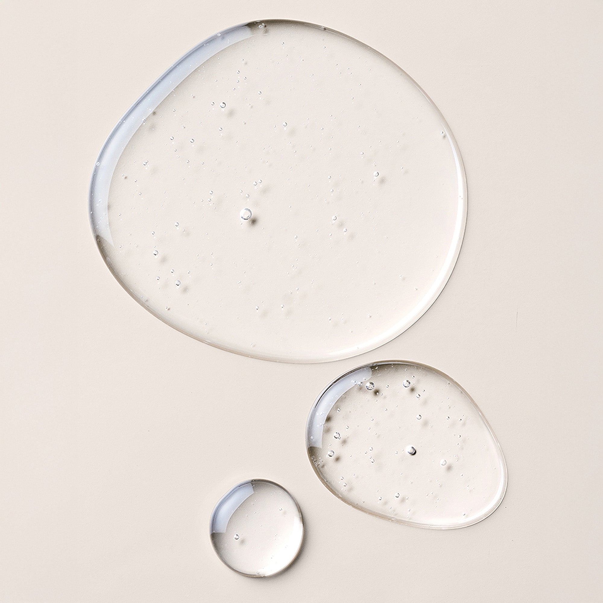 Three droplets of clear, glossy cosmetic gel or serum arranged on a warm beige background, with the largest droplet showing subtle bubbles and a smooth reflective surface.10