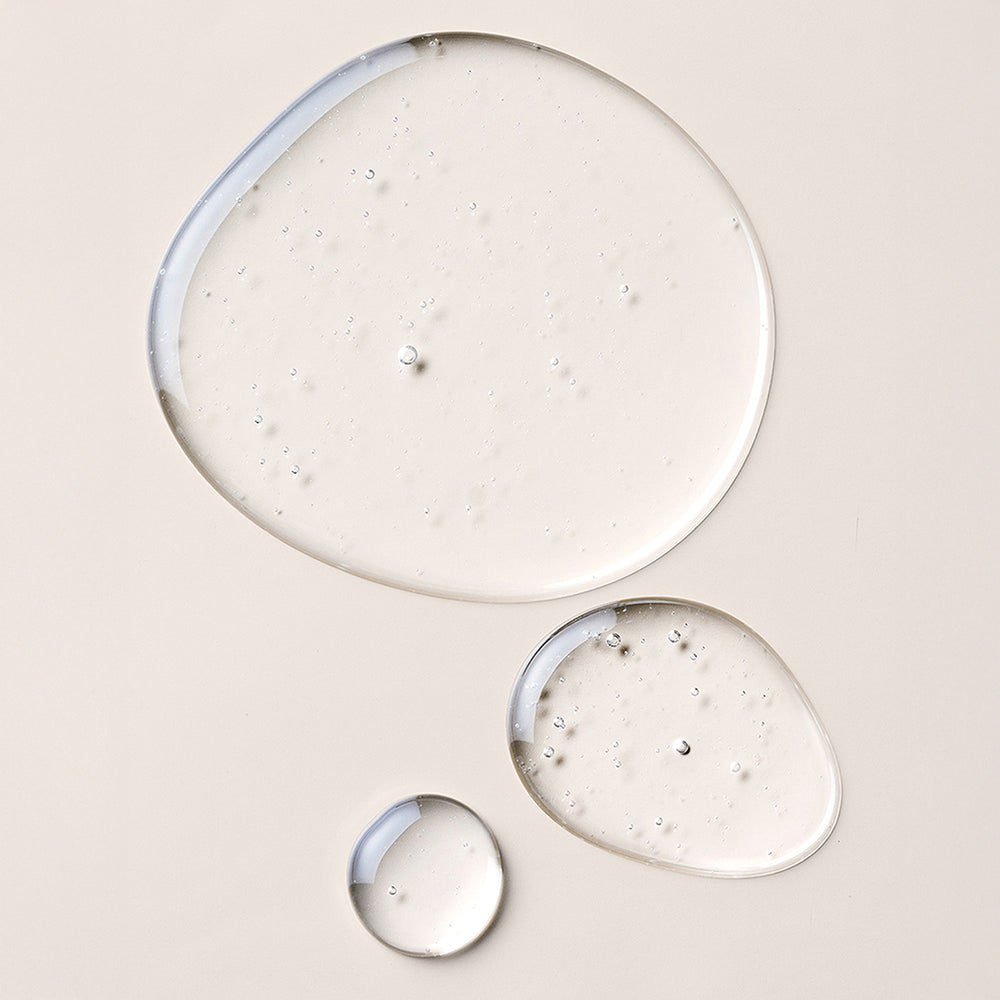 Three droplets of clear, glossy cosmetic gel or serum arranged on a warm beige background, with the largest droplet showing subtle bubbles and a smooth reflective surface.10