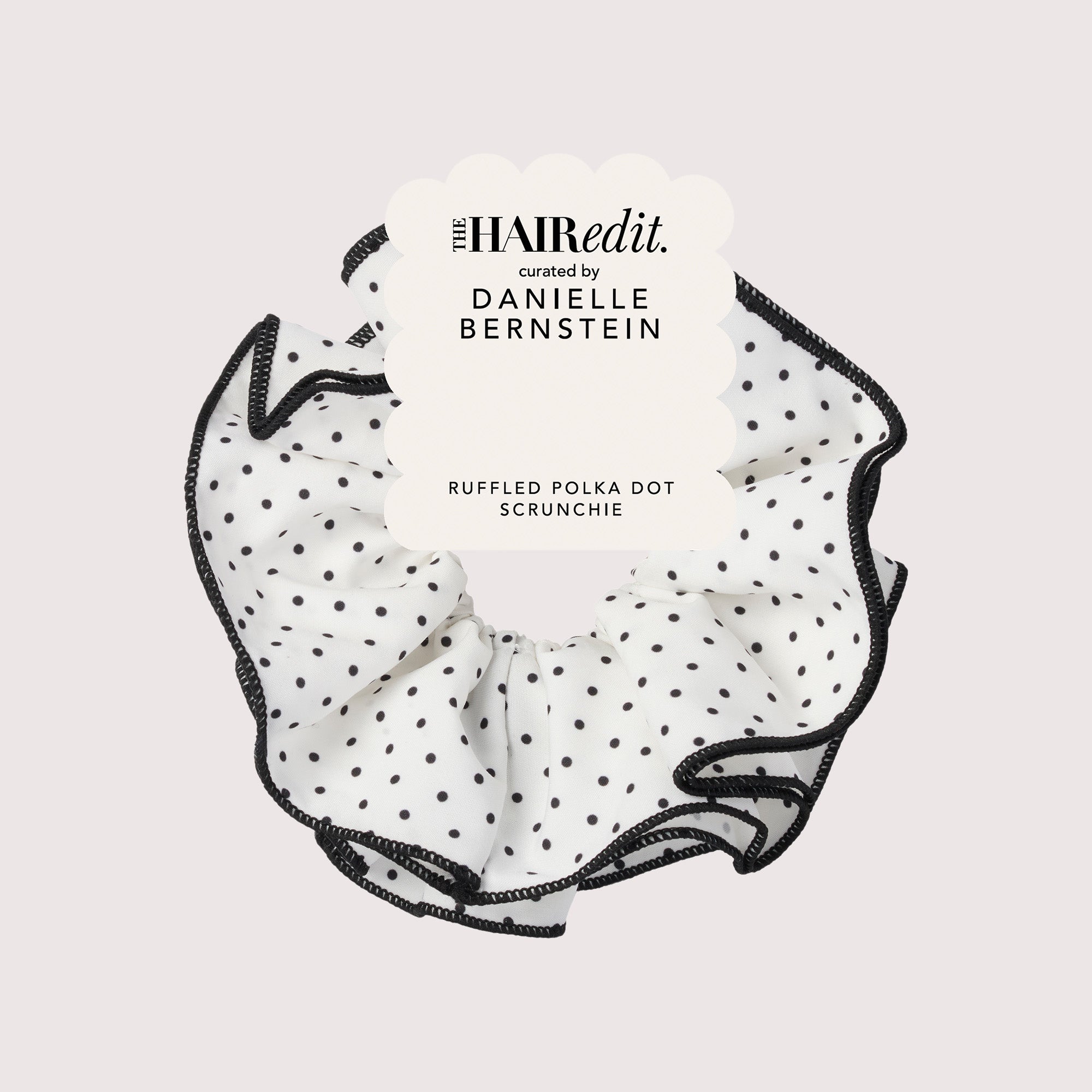 A ruffled polka dot scrunchie by The HAIR EDIT, featuring a white fabric body with black polka dots and black ruffled trim, presented with a white paper tag on a light gray background.11