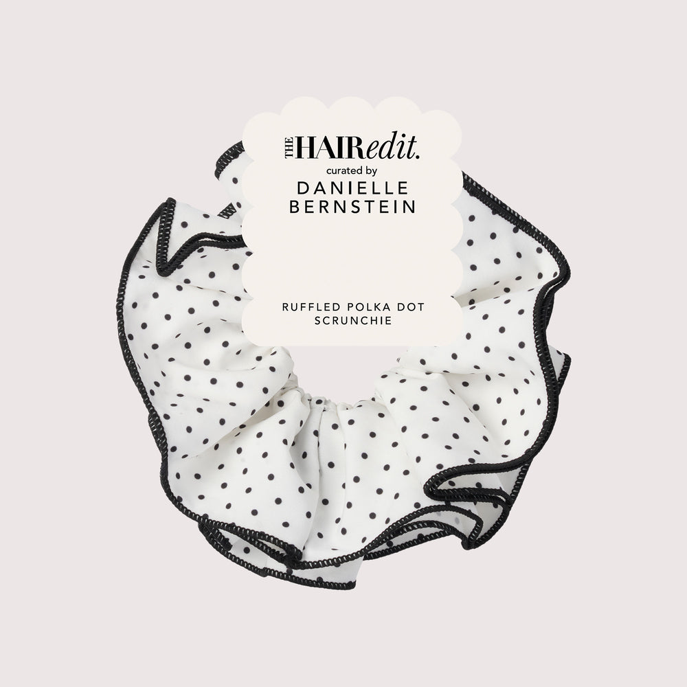 A ruffled polka dot scrunchie by The HAIR EDIT, featuring a white fabric body with black polka dots and black ruffled trim, presented with a white paper tag on a light gray background.11