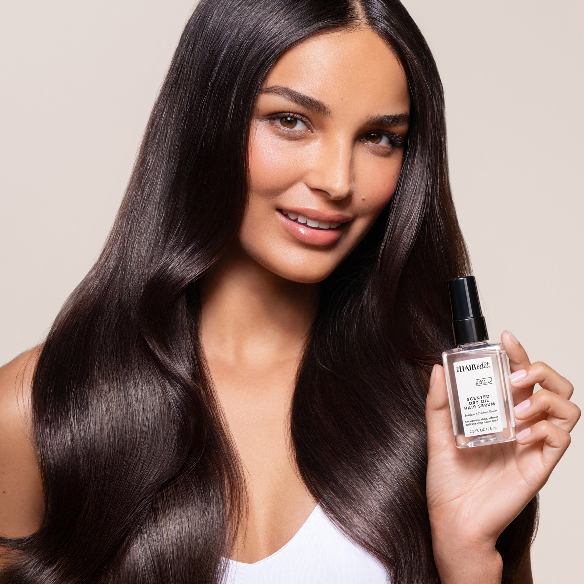 A woman with long, straight, dark brown hair holds a bottle of The Hair Edit Hair Oil in her hand, set against a solid light beige background.8