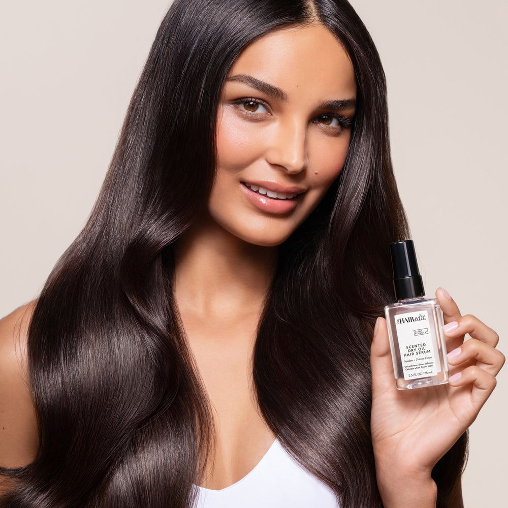 A woman with long, straight, dark brown hair holds a bottle of The Hair Edit Hair Oil in her hand, set against a solid light beige background.8