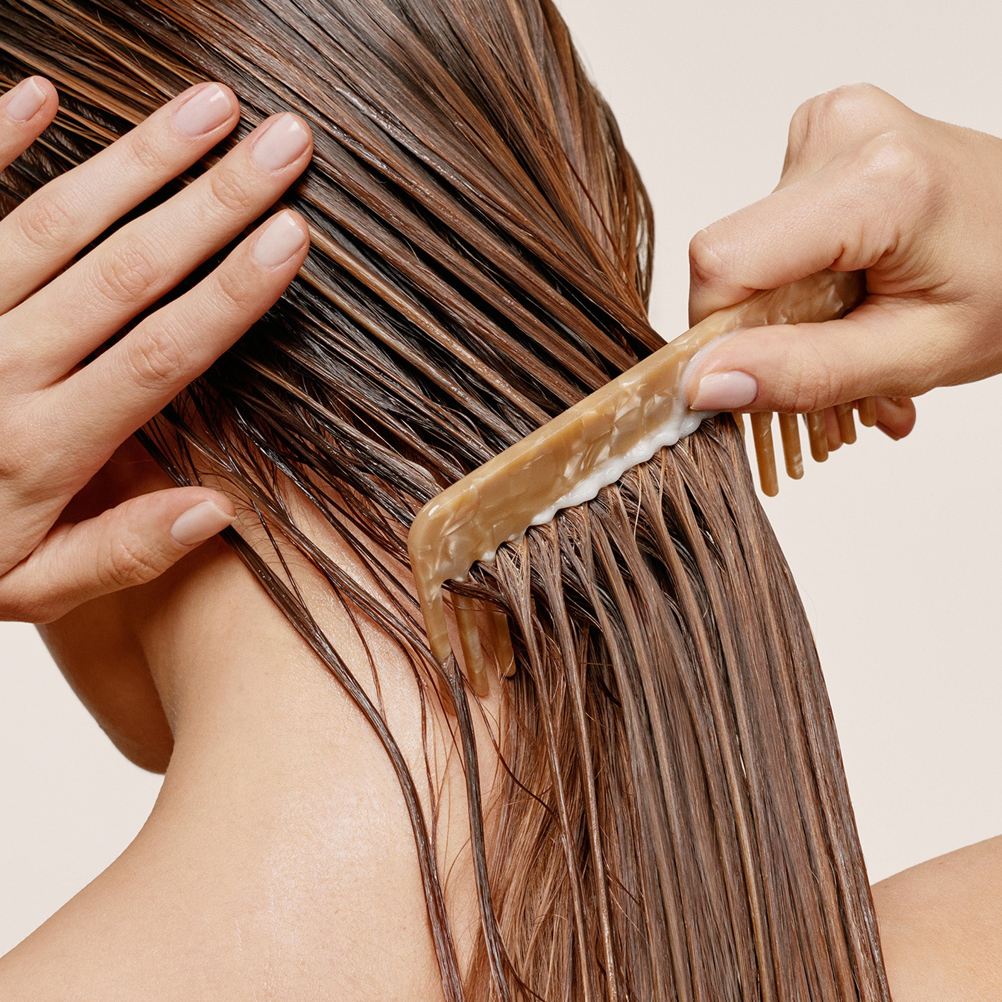 A close-up of a person's wet brown hair being gently combed with a light brown comb, with hands visible against a plain beige background.8