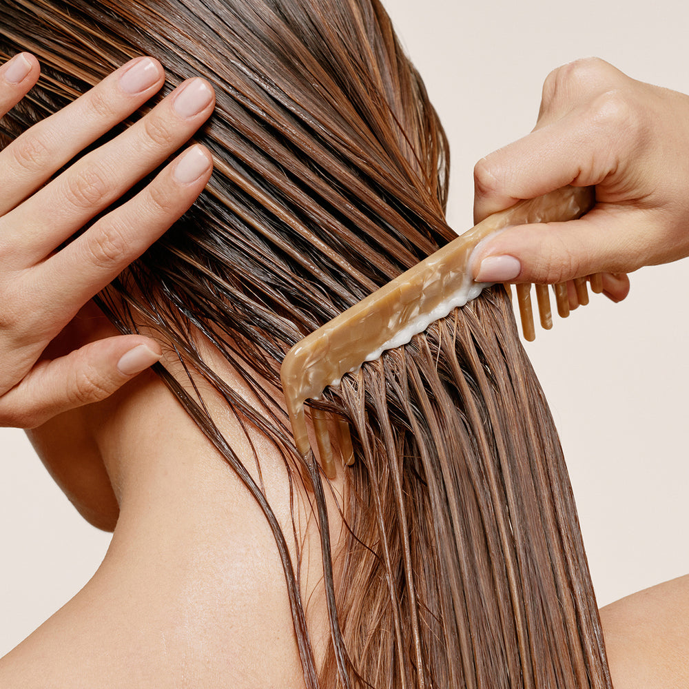 A close-up of a person's wet brown hair being gently combed with a light brown comb, with hands visible against a plain beige background.8