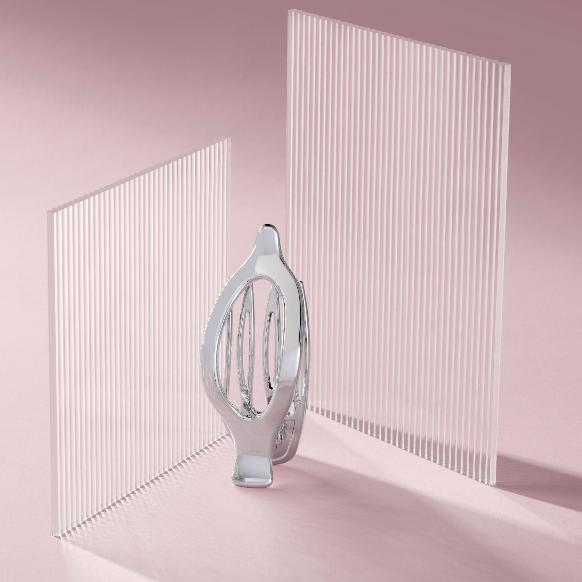 A polished silver metallic flat lay claw clip stands in the center between two tall, translucent white slatted panels on a soft pink studio background.9