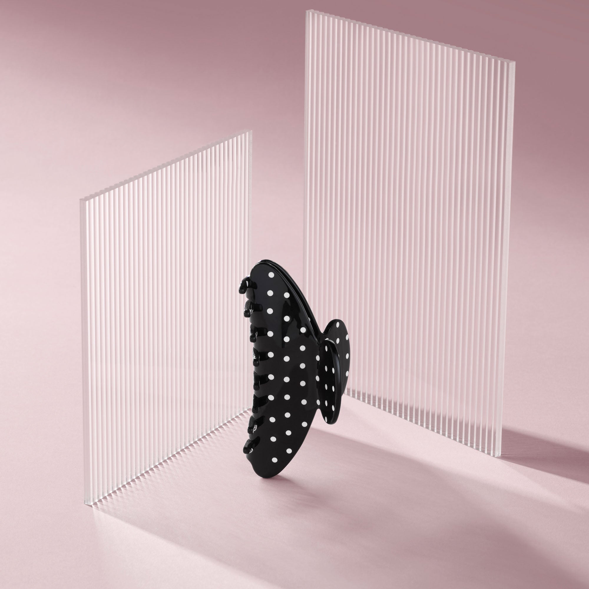 A black hair claw clip with white polka dots, shown floating in front of two translucent white slatted panels on a soft pink studio background.9