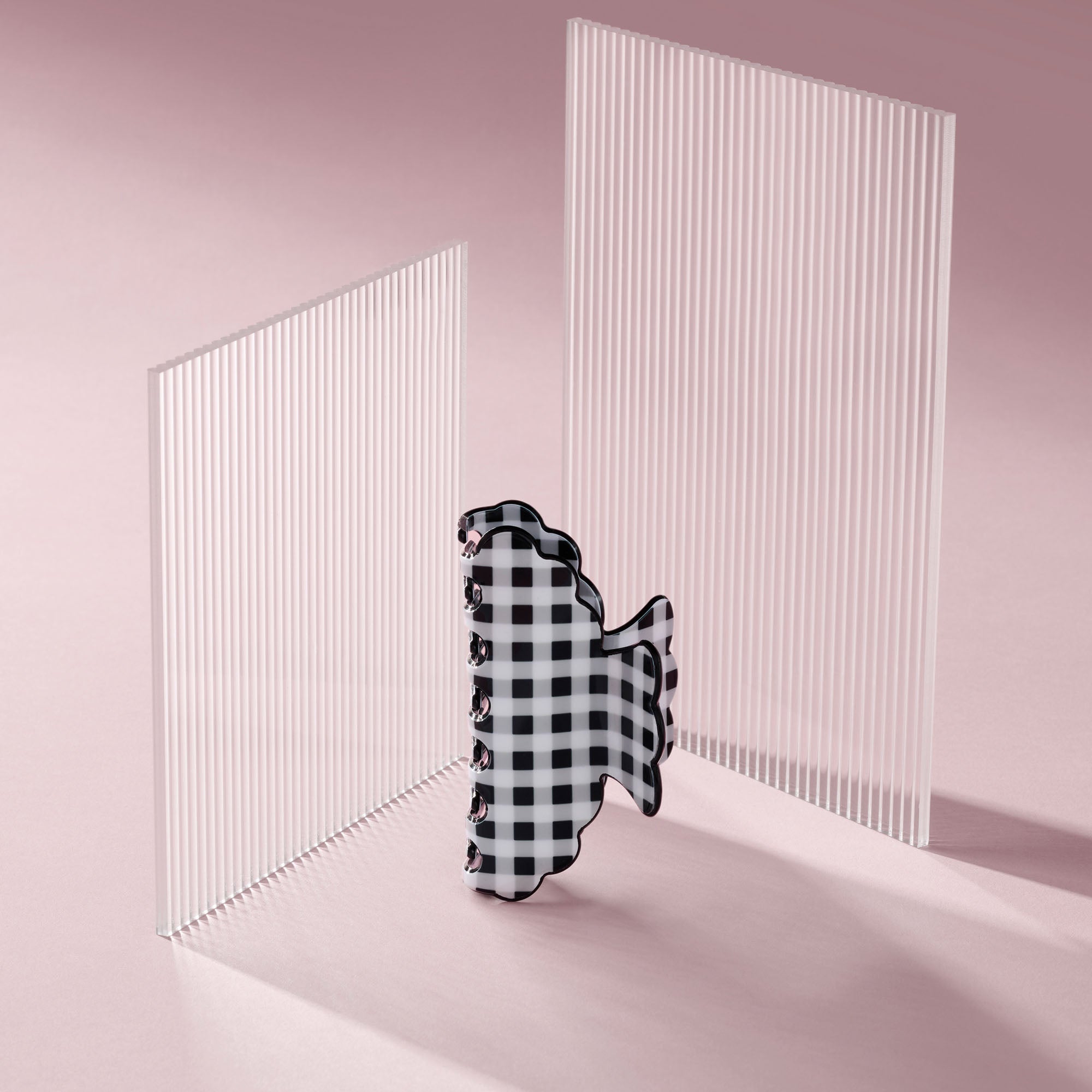 A black and white gingham hair claw clip stands upright between two translucent white acrylic slatted panels on a soft pink studio background.9