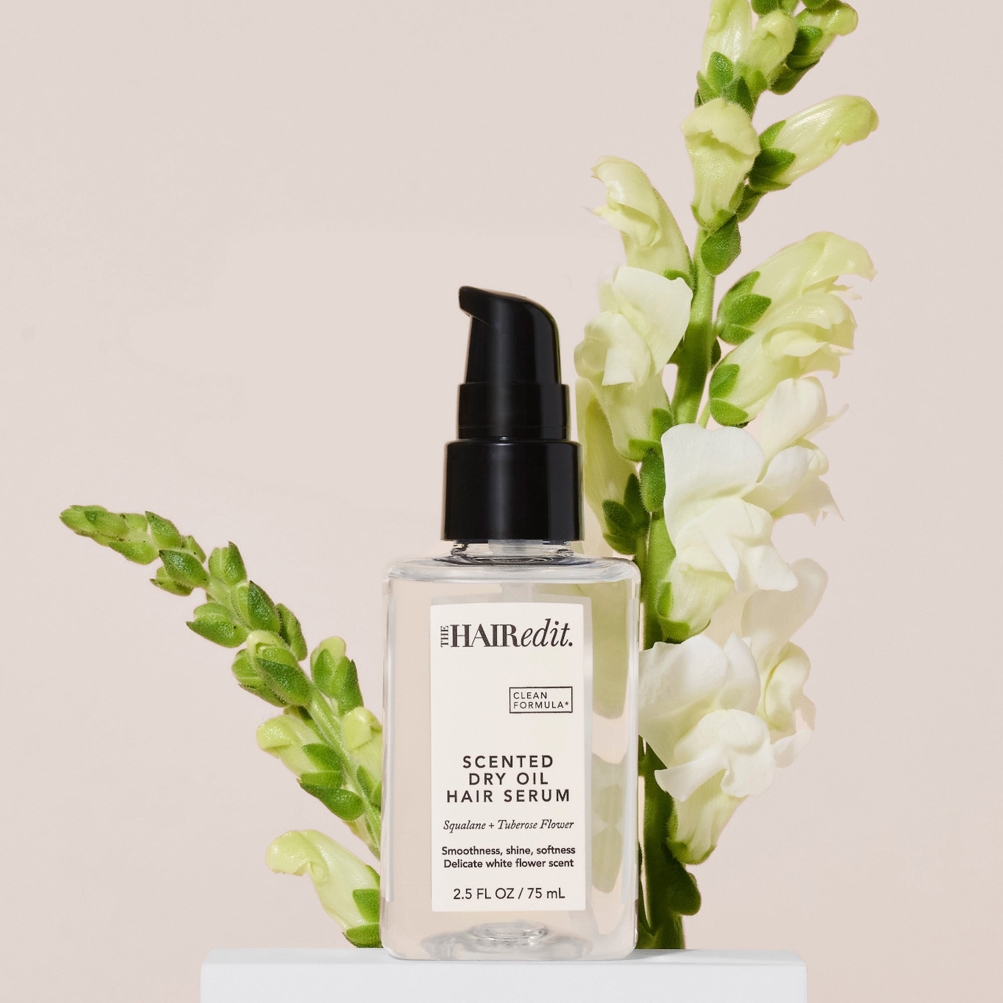 The Hair Edit Scented Dry Oil Hair Serum in a clear bottle with a black pump, styled with white flowers on a light beige background.9